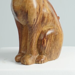 May include: A carved wooden figurine of a sitting cat with a smooth, polished finish. The wood displays natural grain patterns in shades of light and dark brown, highlighting the feline form with visible front paws and a curved back profile.