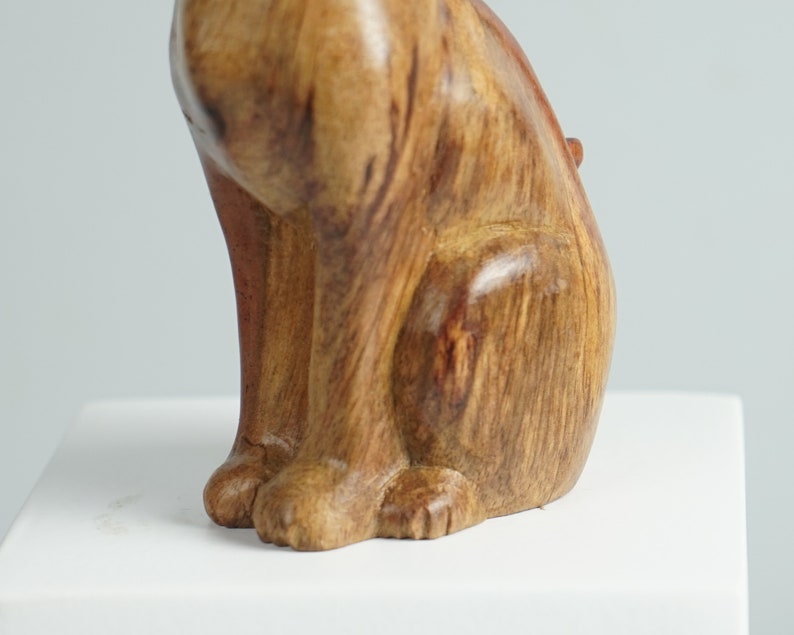 May include: A carved wooden figurine of a sitting cat with a smooth, polished finish. The wood displays natural grain patterns in shades of light and dark brown, highlighting the feline form with visible front paws and a curved back profile.