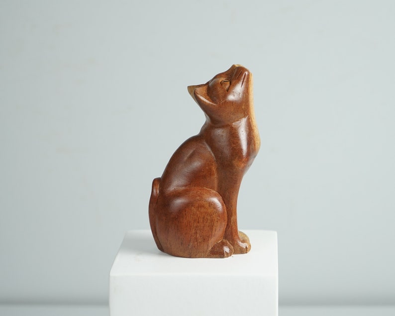 May include: A hand-carved wooden cat figurine in a seated position with its head tilted upward. The sculpture features a smooth, polished medium-brown wood grain finish, showing the natural texture of the material throughout the cat's body.