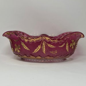 May include: A ruby-coloured glass bowl with a scalloped rim and a pedestal base. The bowl is decorated with gold floral and leaf patterns. The decorative bowl is likely vintage and suitable for display or serving.