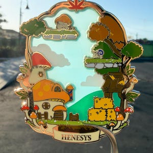 Puede incluir: Un colorido pin de esmalte con una escena de pueblo de hongos caprichosa. El pin incluye una casa de hongos, árboles y el texto "HENESYS" en una pancarta. El diseño tiene un borde dorado y está sujeto por un pasador plateado.