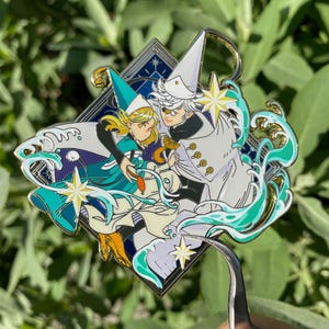 Puede incluir: Un pin de esmalte dorado con dos personajes, uno con un atuendo blanco y azul y el otro con un atuendo blanco, sobre un fondo azul y blanco. El pin tiene forma de diamante y presenta estrellas y olas.