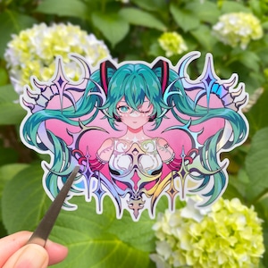 May include: A colorful sticker featuring a stylized anime character with teal hair, winking, and wearing a detailed outfit. The sticker has a pink and white background with decorative elements. The sticker is being held by a pair of tweezers.