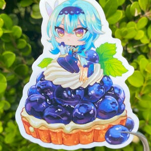 May include: A sticker featuring a cartoon character with blue hair and a blue outfit, sitting atop a blueberry tart. The tart has a golden crust, white cream, and is covered in plump, dark blue blueberries. A green leaf is visible.