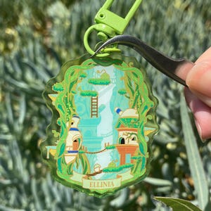 Puede incluir: Un llavero acrílico transparente con un cierre verde. El llavero presenta una ilustración caprichosa de un paisaje de fantasía con edificios, escaleras y la palabra "ELLINIA" en una pancarta. El diseño está delineado en dorado.