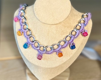 Könnte beinhalten: Eine lila gehäkelte Halskette mit einer silbernen Kette und bunten Edelsteinperlen. Die Halskette wird auf einer Puppe präsentiert.