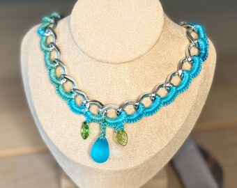Könnte beinhalten: Eine blaue und grüne gehäkelte Halskette mit einer silbernen Kette und einem blauen Glasanhänger. Die Halskette befindet sich auf einem Präsentationsständer.
