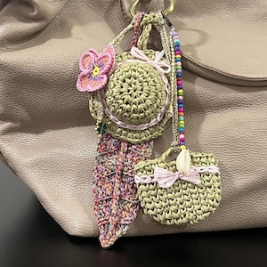 May include: A handmade crochet bag charm featuring a small hat with a pink flower, a leaf, and a small purse. The charm is attached to a beige leather bag. The charm has pink and green accents.