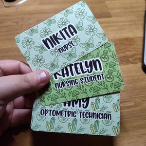 May include: Three personalized name badges with unique designs and professions. The top badge reads "NIKITA nurse" with a floral and butterfly pattern. The middle badge says "KATELYN nursing student" with a botanical theme. The bottom badge reads "AMY Optometric Technician" with a crystal design.