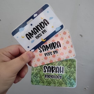 May include: Three personalized name badges with different designs. The top badge has a blue border with the name "AMANDA" and "NICU RN" on a background of clouds and stars. The middle badge has the name "SAMIRA" and "PEDS RN" on a peach-colored background. The bottom badge has the name "SARAH" and "RADIOLOGY" on a green background.
