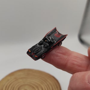 Puede incluir: Una Batmobile negra en miniatura con detalles rojos, sentada en un dedo.