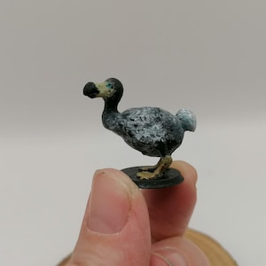 Puede incluir: Una pequeña figurita pintada a mano de un dodo. El dodo es gris con manchas blancas y tiene un pico negro y ojos azules. La figurita está de pie sobre una base negra.