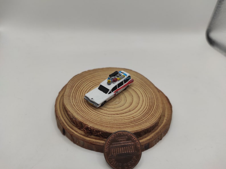 May include: A miniature white Ghostbusters car with red and blue accents on a wooden base. The car has a roof rack with several items on it.