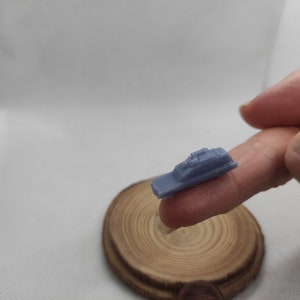 May include: A small, light blue, 3D printed model of a police car. The model is being held by a finger on a wooden surface.