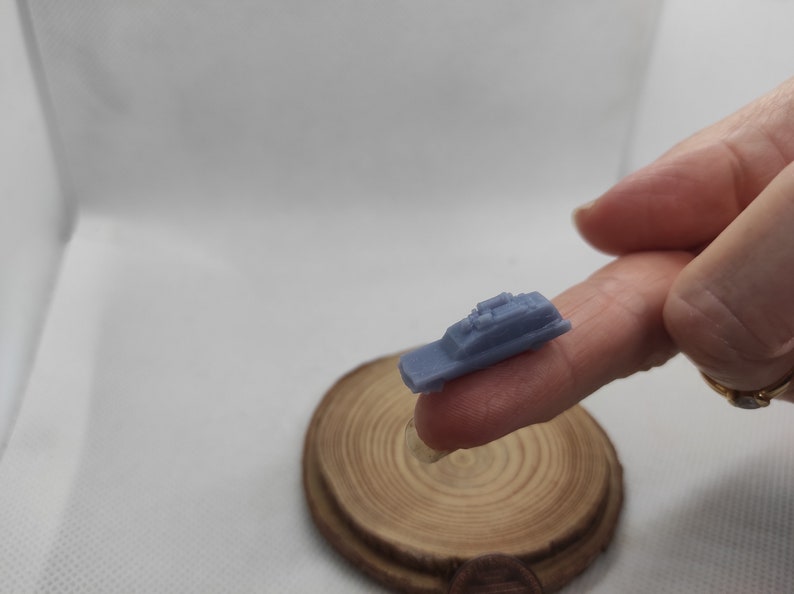 May include: A small, light blue, 3D printed model of a police car. The model is being held by a finger on a wooden surface.