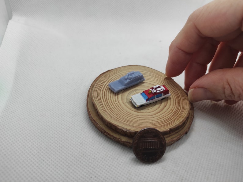 May include: Two miniature toy cars on a wooden base. The car on the left is a light blue and white police car. The car on the right is a red and white ambulance. A penny is in the foreground.