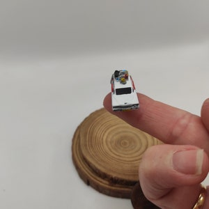 May include: A miniature white car with red accents and a ghostbusters logo on the side. The car has a small figure on top and is sitting on a finger.