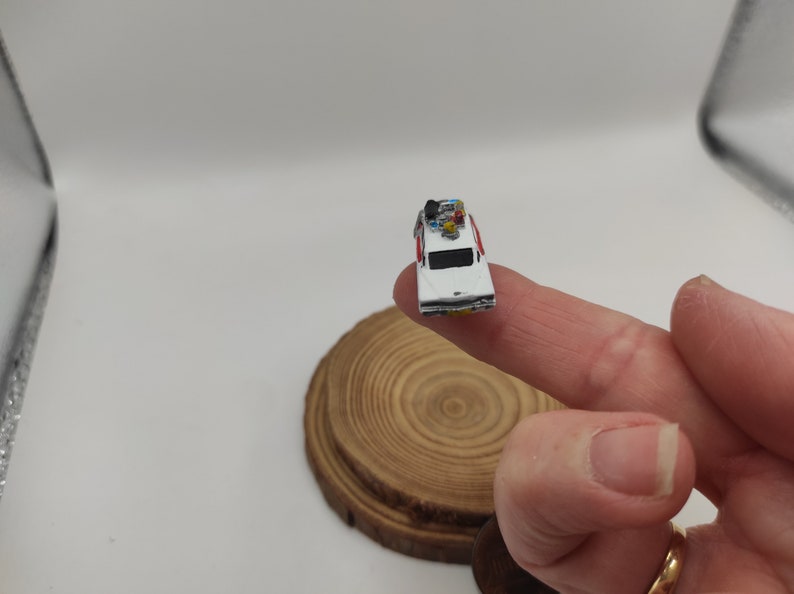 May include: A miniature white car with red accents and a ghostbusters logo on the side. The car has a small figure on top and is sitting on a finger.