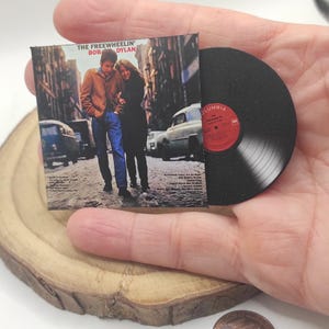 Peut inclure: Miniature de la pochette de l'album "The Freewheelin'" de Bob Dylan et d'un disque vinyle. La pochette présente une photo en noir et blanc d'un couple marchant dans une rue. Le disque est noir avec une étiquette rouge.
