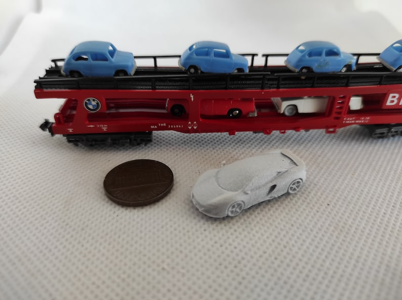Puede incluir: Un vag&oacute;n de tren rojo de juguete con un marco de metal negro y un coche blanco en el nivel superior. El vag&oacute;n lleva cuatro coches de juguete azules. El vag&oacute;n tiene el texto "THE" y "MA 335611" impreso en el lateral. Un coche de juguete blanco est&aacute; en el suelo delante del vag&oacute;n.