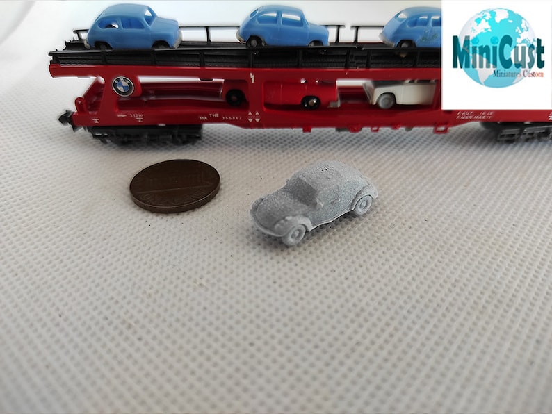 Puede incluir: Un coche modelo gris sobre una superficie blanca. El coche es un Volkswagen Beetle cl&aacute;sico de forma redondeada y tama&ntilde;o peque&ntilde;o. El coche est&aacute; sobre una superficie blanca con un patr&oacute;n texturizado. El coche es un modelo en miniatura y tiene aproximadamente el tama&ntilde;o de una moneda de un centavo.