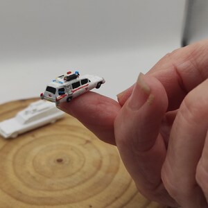 May include: A miniature white Ghostbusters car with red and blue details, sitting on a finger. The car is a replica of the iconic Ecto-1 vehicle from the Ghostbusters movie.