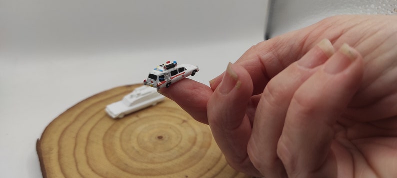 May include: A miniature white Ghostbusters car with red and blue details, sitting on a finger. The car is a replica of the iconic Ecto-1 vehicle from the Ghostbusters movie.