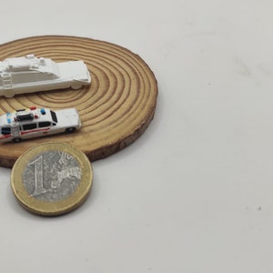 May include: Two miniature white toy cars on a wooden base. The car on the left is a police car with red and blue lights. The car on the right is a van with a ladder on top. A euro coin is in the foreground.