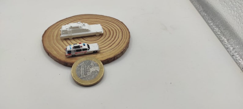 May include: Two miniature white toy cars on a wooden base. The car on the left is a police car with red and blue lights. The car on the right is a van with a ladder on top. A euro coin is in the foreground.