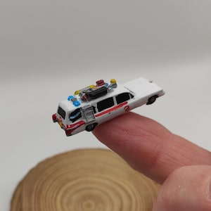 May include: A miniature white and red Ghostbusters car with a ladder on the side and a black box on the roof. The car has blue and yellow lights on top.