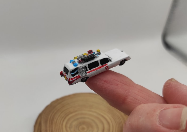 May include: A miniature white and red Ghostbusters car with a ladder on the side and a black box on the roof. The car has blue and yellow lights on top.