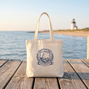 Könnte beinhalten: Eine naturfarbene Stofftasche aus Segeltuch mit einem blauen Grafikdruck eines Leuchtturms in einem Wappen. Der Text lautet NANTUCKET MASSACHUSETTS EST. 1829. Die Tasche hat zwei Schulterriemen und einen rechteckigen Körper mit flachem Boden.