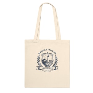 Könnte beinhalten: Eine naturfarbene Stofftasche aus Segeltuch mit einem dunkelblauen Grafikdruck. Das Motiv zeigt einen Leuchtturm und ein Gebäude in einem Wappen, flankiert von Lorbeerzweigen, mit dem Text Martha's Vineyard, Massachusetts und Est. 1603.