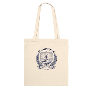 May include: A cream-coloured canvas tote bag featuring a dark blue graphic print. The design includes a shield with a windmill, surrounded by laurel leaves, with the text HAMPTONS NEW YORK and EST. 1640 printed in a clear, dark blue font.
