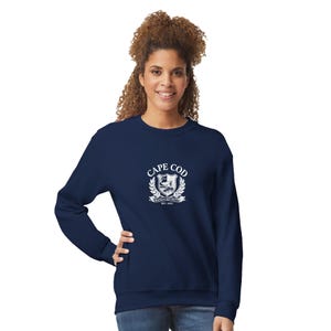 Könnte beinhalten: Ein dunkelblaues Langarm-Sweatshirt mit einem weißen Grafikdruck auf der Brust. Das Design zeigt ein Wappen mit einem Wal, umgeben von Lorbeerblättern, sowie den darunter gedruckten Text CAPE COD, MASSACHUSETTS und EST. 1602.