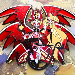 Op de afbeelding: Een gedetailleerde emaille pin met twee gestileerde figuren met rode vleugels en hoorns. De figuren zijn rood, zwart, wit en geel. E&eacute;n figuur houdt een vlam vast. De pin heeft een zwarte omtrek en een glanzende afwerking.