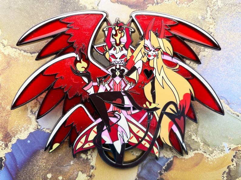 Op de afbeelding: Een gedetailleerde emaille pin met twee gestileerde figuren met rode vleugels en hoorns. De figuren zijn rood, zwart, wit en geel. E&eacute;n figuur houdt een vlam vast. De pin heeft een zwarte omtrek en een glanzende afwerking.