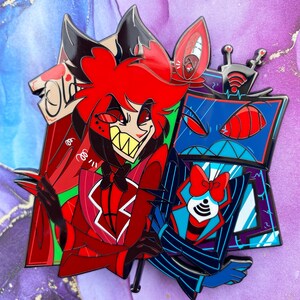 Op de afbeelding: Een kleurrijke emaille pin met twee stripfiguren. De ene is rood met een brede grijns, de andere is blauw met een tv-schermhoofd. De pin heeft een zwarte omtrek en staat tegen een paarse en witte achtergrond.