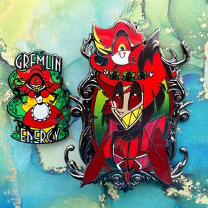 Op de afbeelding: Twee emaille pins met stripfiguren. E&eacute;n pin heeft een rood personage met de tekst "GREMLIN ENERGY" in het groen. De andere pin heeft een rood personage in een zwart, sierlijk frame.