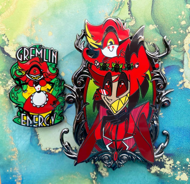 Op de afbeelding: Twee emaille pins met stripfiguren. E&eacute;n pin heeft een rood personage met de tekst "GREMLIN ENERGY" in het groen. De andere pin heeft een rood personage in een zwart, sierlijk frame.