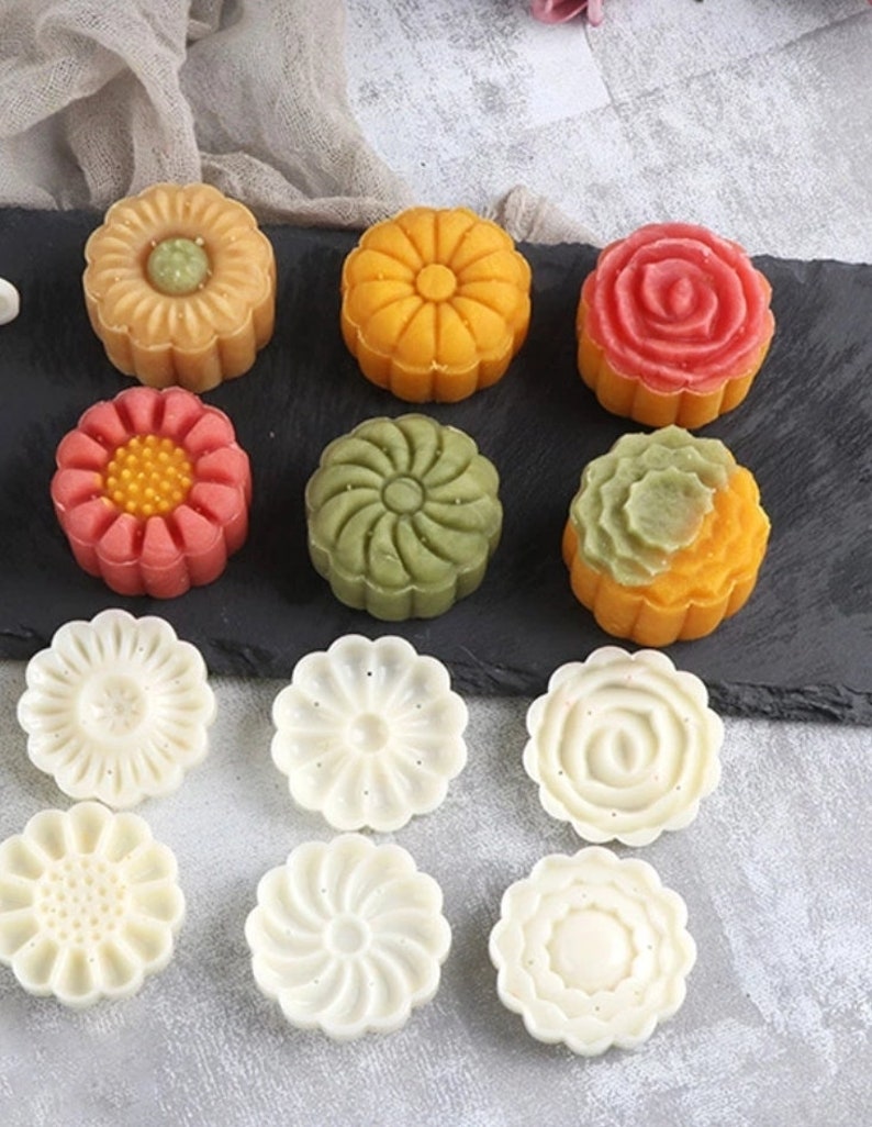 Pode incluir: Conjunto de mooncakes coloridos com v&aacute;rios desenhos de flores, incluindo uma margarida amarela, uma rosa rosa, um redemoinho verde e uma flor vermelha. Tamb&eacute;m est&atilde;o inclu&iacute;dos seis moldes de pl&aacute;stico branco com desenhos de flores.