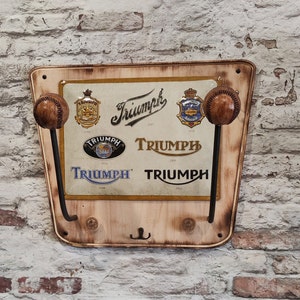 Op de afbeelding: Houten wandkapstok met Triumph-logo. Voorzien van twee honkbalvormige knoppen en diverse Triumph-logo's uit verschillende jaren. De kapstok heeft een verweerde uitstraling, met een lichtbruine afwerking en donkere metalen haken.