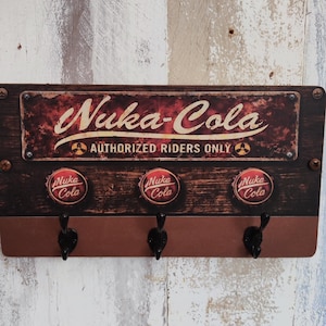 Może przedstawiać: Drewniany wieszak na klucze do montażu na ścianie z postarzanym wykończeniem. Znak głosi "Nuka-Cola Authorized Riders Only" w stylu vintage. Posiada trzy haczyki na klucze pod znakiem, z wzorami kapsli Nuka-Cola.