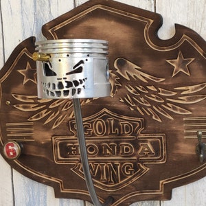 Può includere: Un portachiavi da parete in legno a tema motociclistico. La targa in legno marrone scuro presenta un design intagliato con ali, stelle e le parole "Gold Honda Wing". Un pistone riutilizzato con un design a teschio tiene una chiave. Include un gancio.