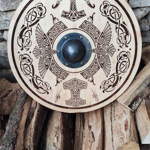 Könnte beinhalten: Ein runder Holzschild mit aufwendigen Wikinger-Gravuren. Das Design zeigt eine zentrale Nabe, Hammelsymbole, Vögel und Knotenmuster. Der Schild ist hellfarben mit dunkelbraunen geätzten Details, vor dem Hintergrund von gestapeltem Brennholz.
