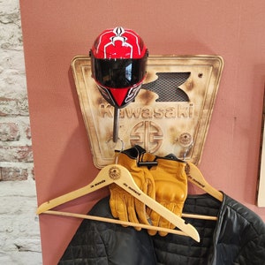 Peut inclure: Un casque de moto rouge et noir est monté au-dessus d'une enseigne Kawasaki en bois. En dessous, une paire de gants en cuir beige sont suspendus à un cintre en bois, avec une veste noire matelassée. Les articles sont exposés sur un mur rose.