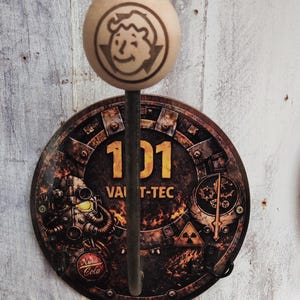 Może przedstawiać: Hak ścienny z motywem Fallout, z drewnianą kulą z wizerunkiem Vault Boya mrugającego okiem. Hak ma okrągłą metalową płytkę z numerem 101, tekstem Vault-Tec i innymi obrazami z Fallout. Kolorystyka to brązowy, czarny i żółty.
