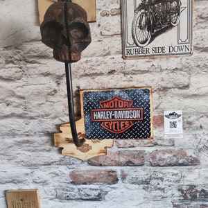 Può includere: Un gancio da parete a forma di teschio in legno con un'asta di metallo, montato su un muro di mattoni. Sono visibili anche insegne Harley-Davidson e un'insegna di moto d'epoca. Le insegne presentano il logo Harley-Davidson e le parole "Rubber Side Down."