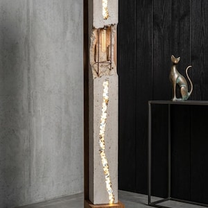 May include: A tall, modern floor lamp with a concrete and wood design. The lamp features a rectangular concrete column with a vertical illuminated strip of embedded stones. The base and top are made of wood. A bronze cat statue sits nearby.