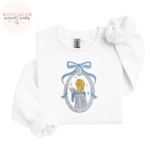 May include: White sweatshirt with an embroidered design. The design features a blue bow, a framed image of a person with blonde hair, and blue clothing. The sweatshirt has a crew neck and long sleeves. The brand name "Gildan" is visible.
